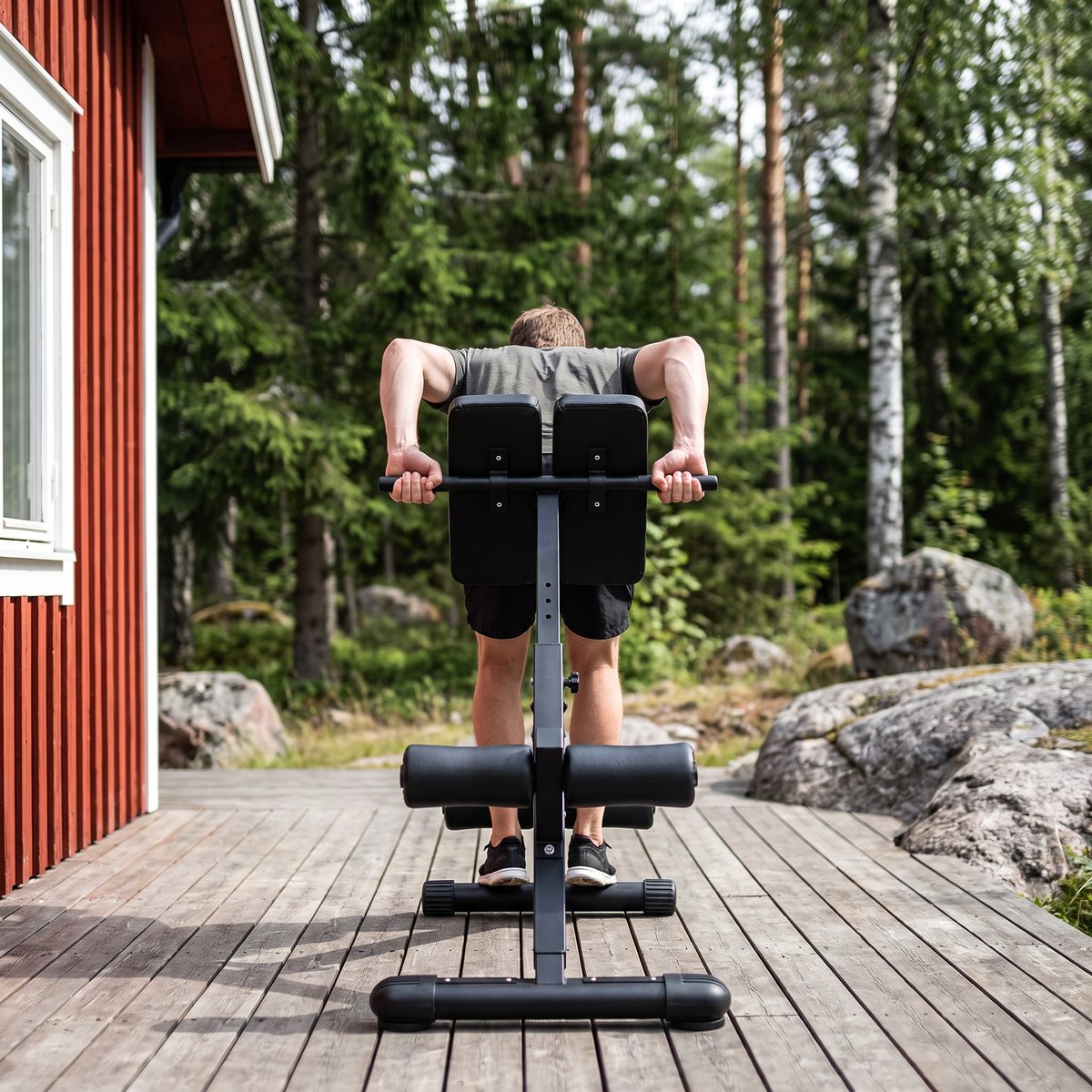 Person gör rygglyft på en svart hopfällbar ryggbänk på en trätrall vid en röd stuga i svensk skog