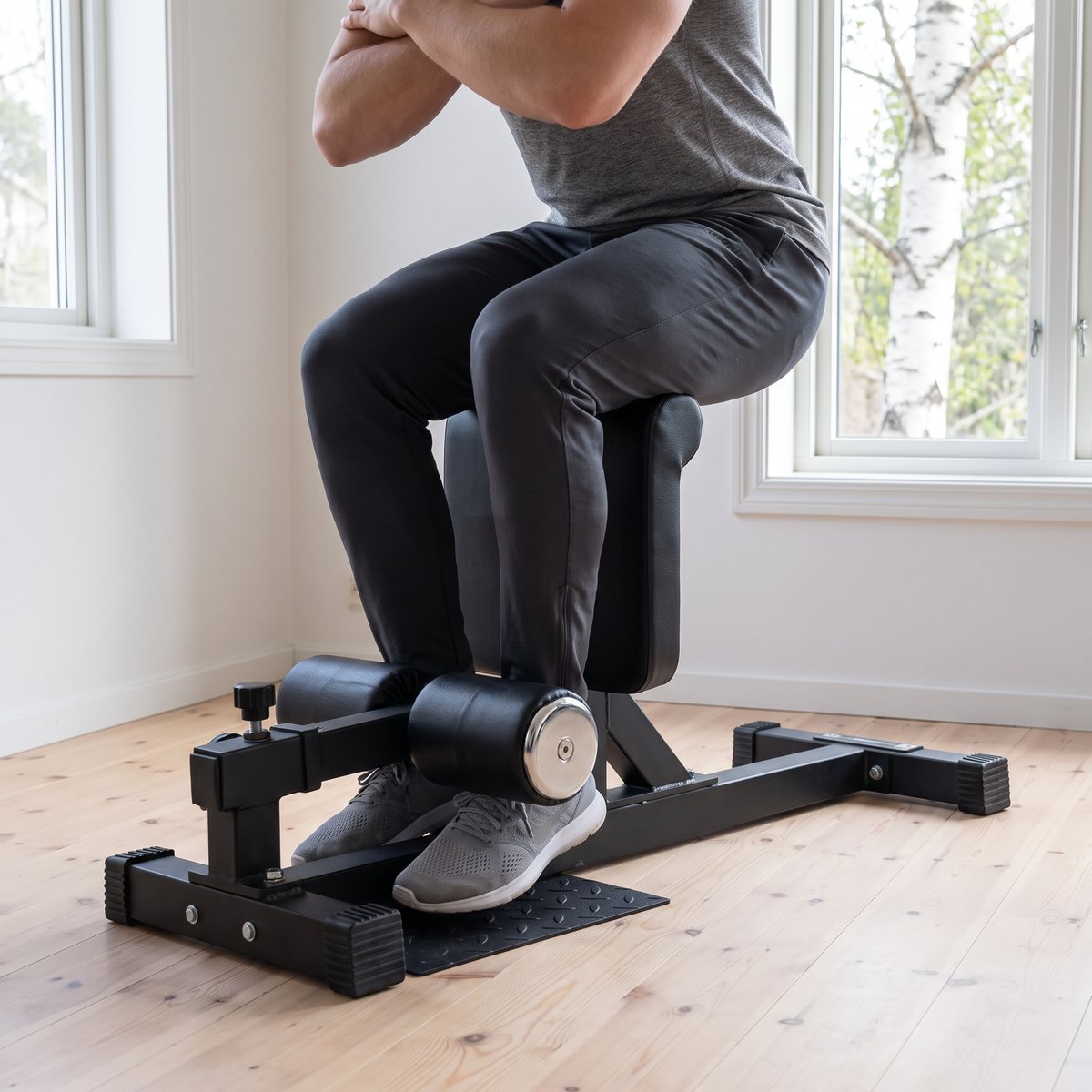Sissy squat-bänken inSPORTline ON-X SSB10 i svart stål används i ett ljust svenskt hemmagym med furugolv och björk utanför fönstret