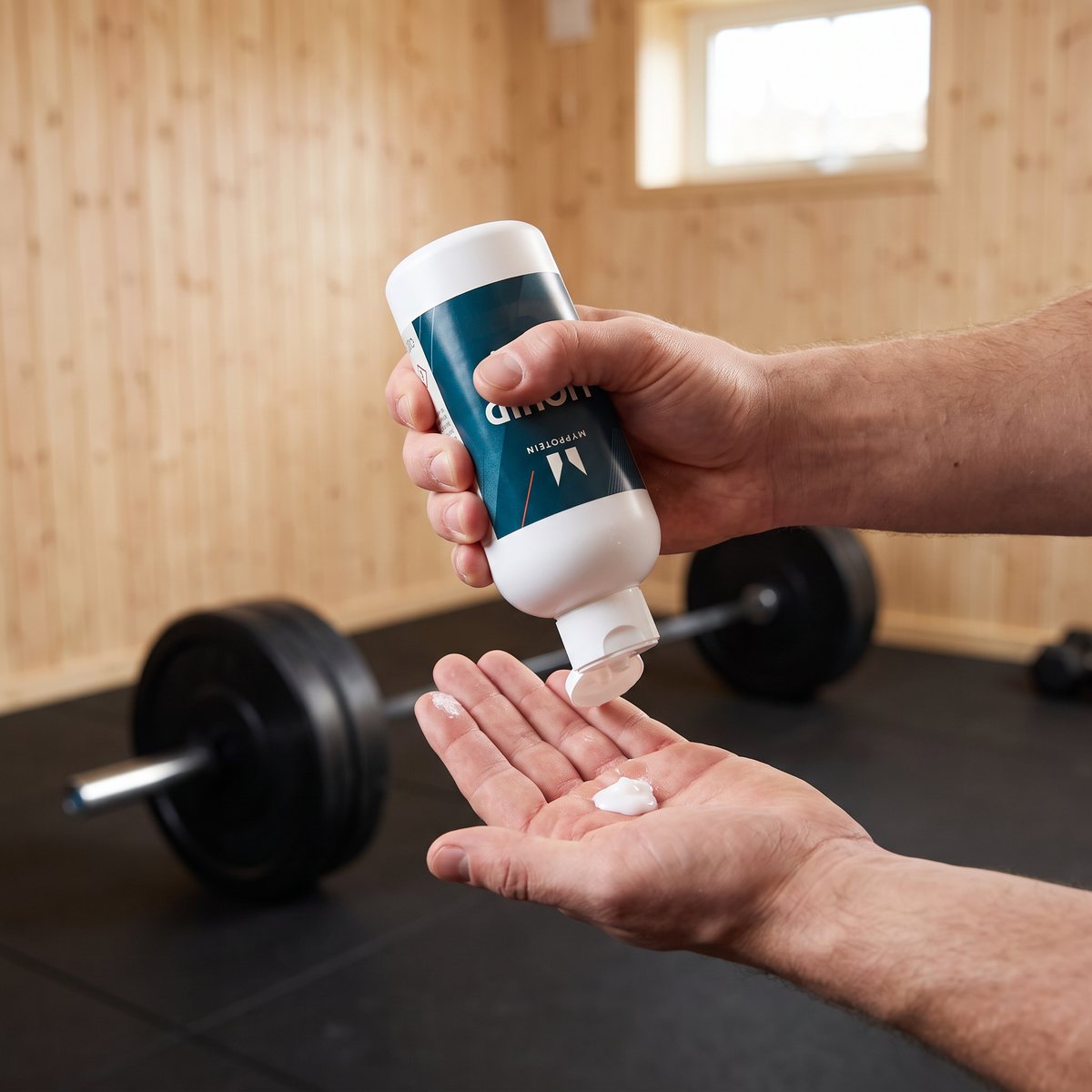Hand som håller en vit flaska MyProtein Liquid Chalk och kramar vätskekritan i andra handflatan i ett svenskt hemmagym med furupanel