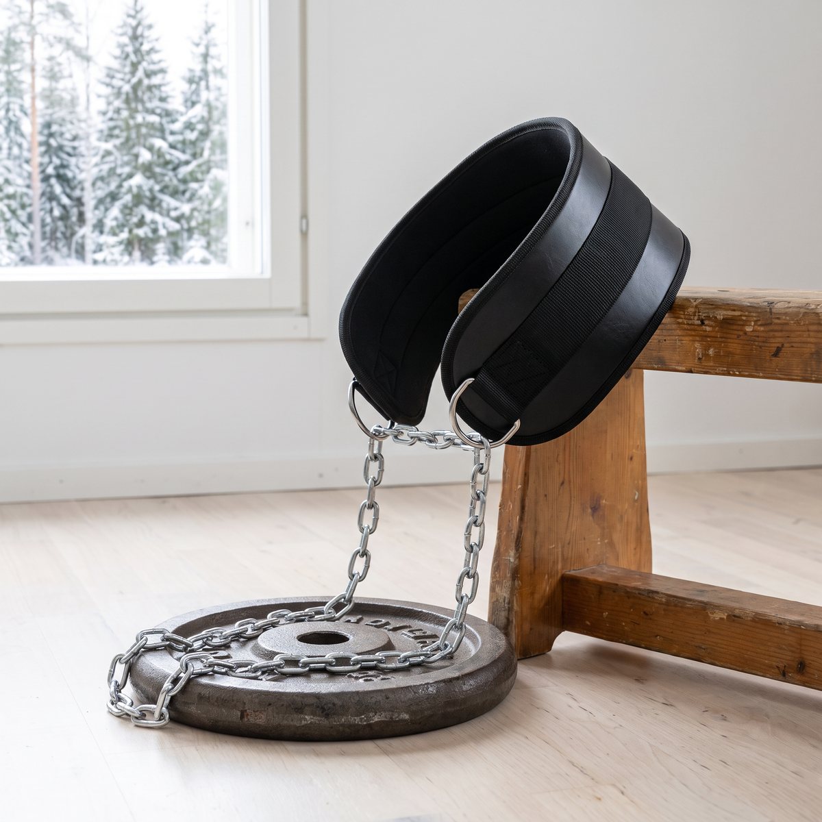 Svart dipbälte med kedja vilande på en träbänk i ett svenskt hemmagym med snötäckta granar utanför fönstret