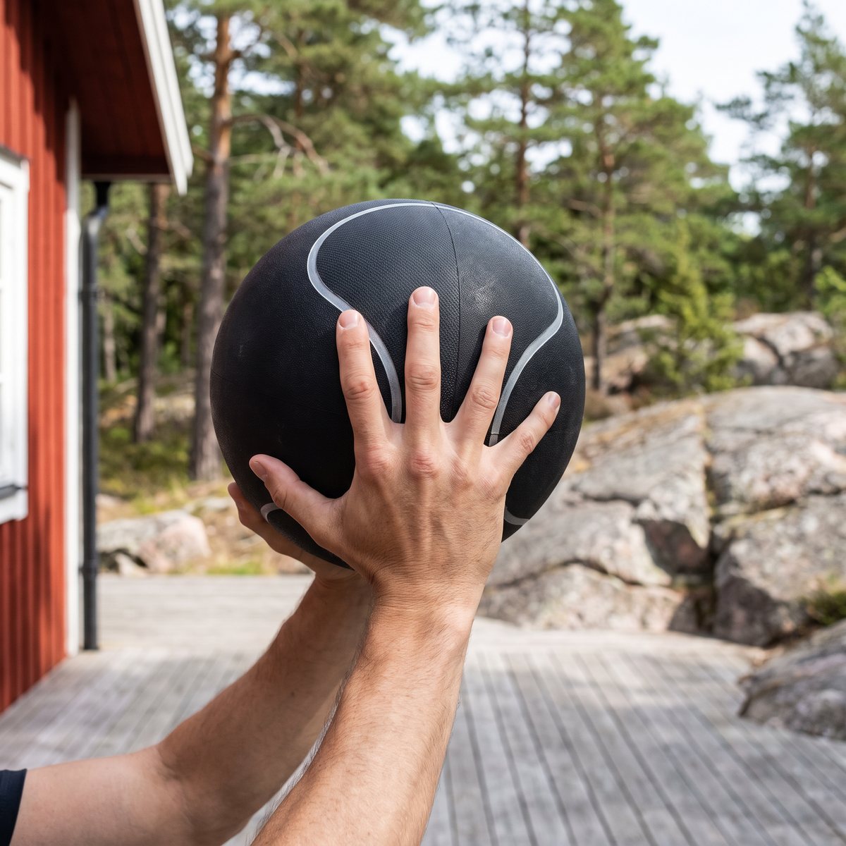 Svart gummimedicinboll med silvergrå sömlinje hålls över huvudet på en trädäck vid en röd stuga med tallar och granit