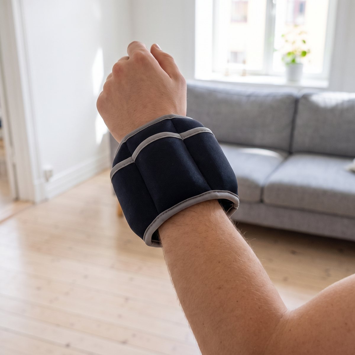 Iron Gyms mörka viktmanschett med grå kant spänd runt handleden i ett svenskt vardagsrum med soffa och ljust trägolv