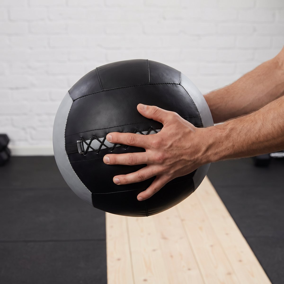 Svart wall ball med grå sidopanel hålls i båda händerna i ett svenskt hemmagym med vitmålad tegelvägg och furugolv