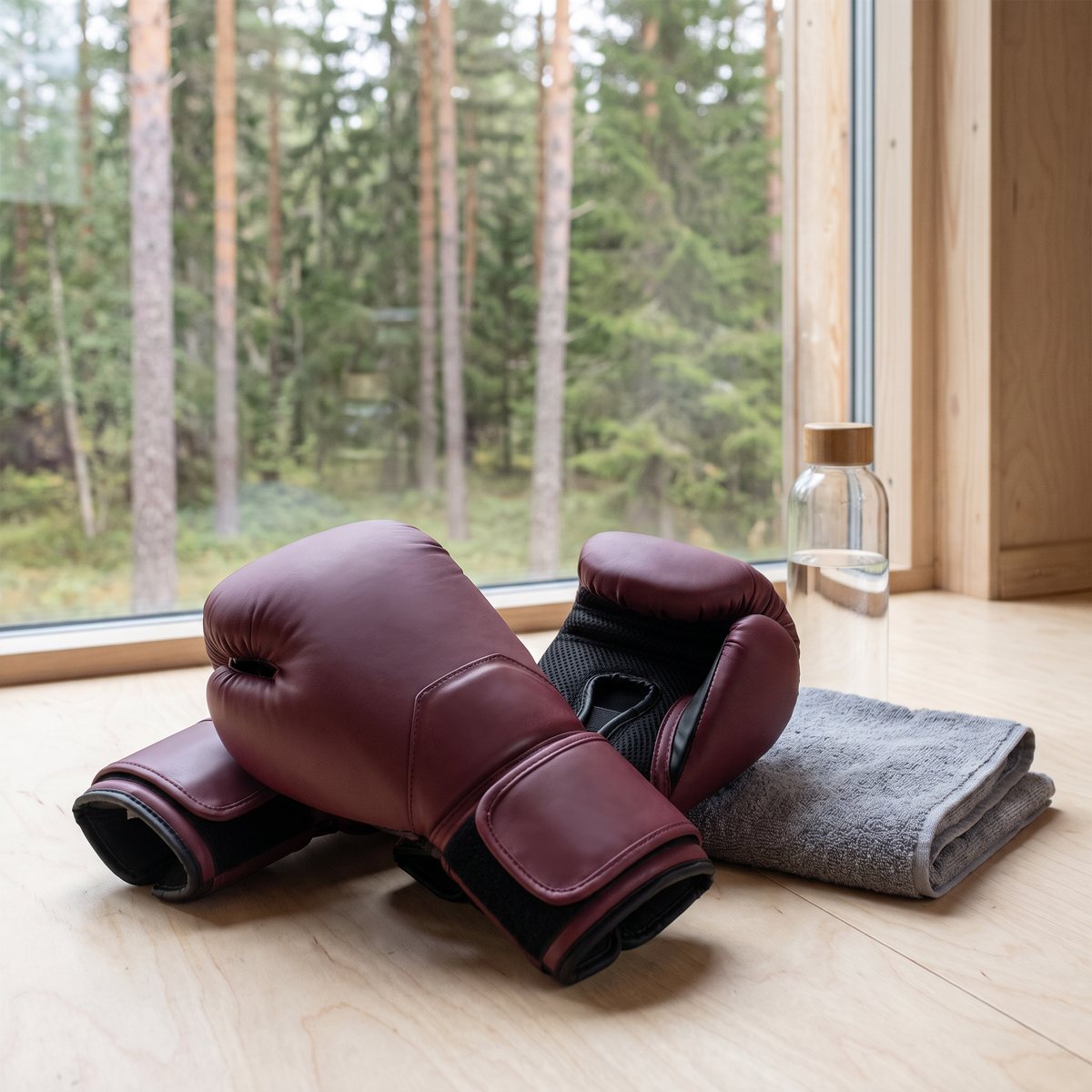 Vinröda Everlast Elite 2-boxningshandskar på ett ljust trägolv bredvid en handduk och en vattenflaska vid ett fönster mot tallskog