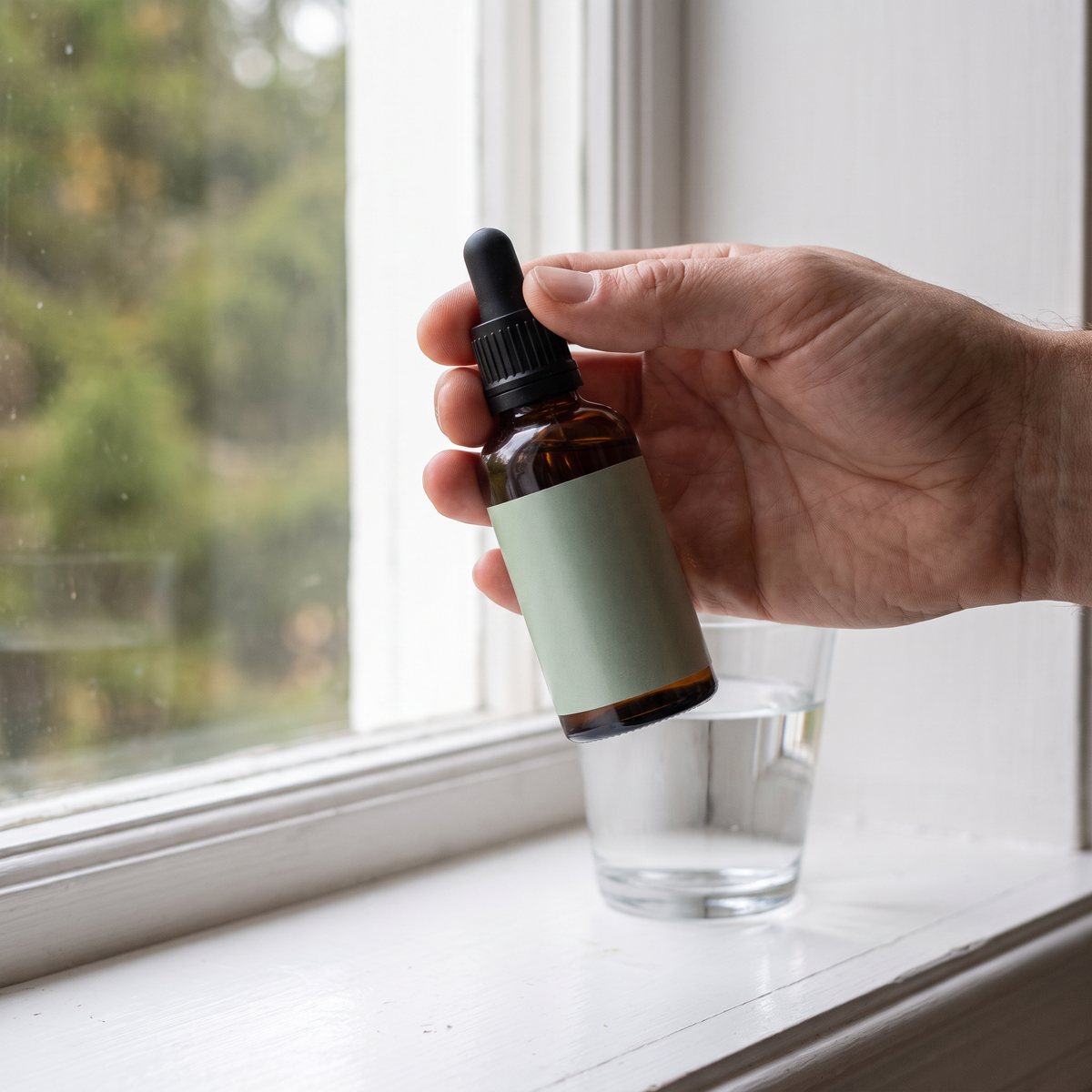 Droppflaska med Örtspecialisten Astragalus i en hand vid ett fönster med glas vatten i ett svenskt hem