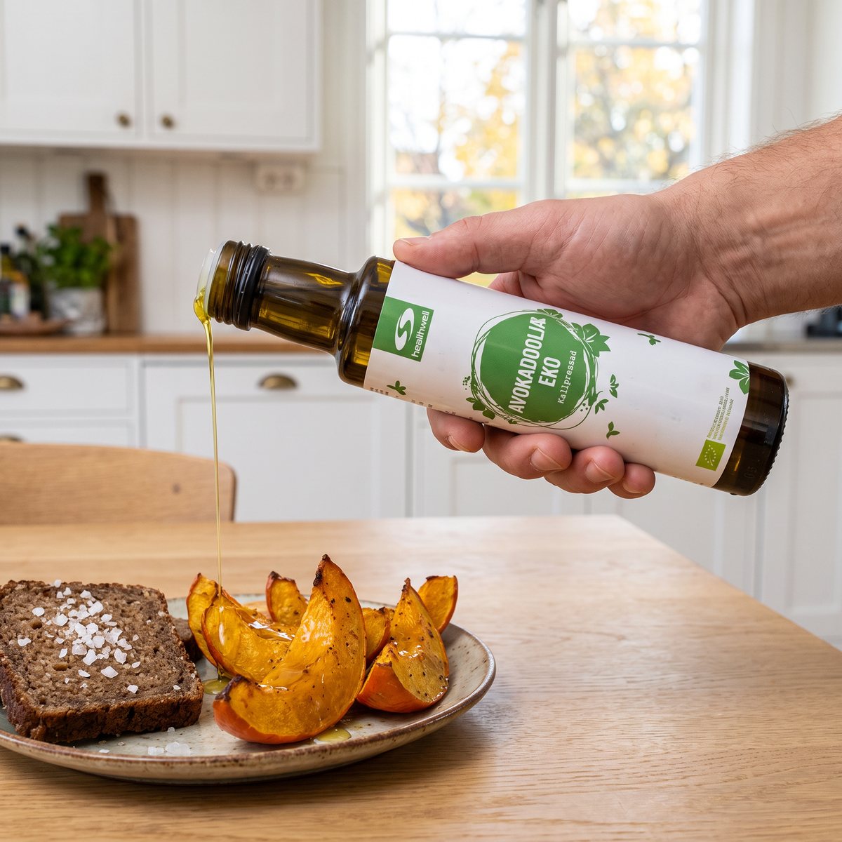 Healthwell Avokadoolja EKO ringlas över ugnsrostad pumpa bredvid en skiva rågbröd med flingsalt i ett ljust svenskt kök