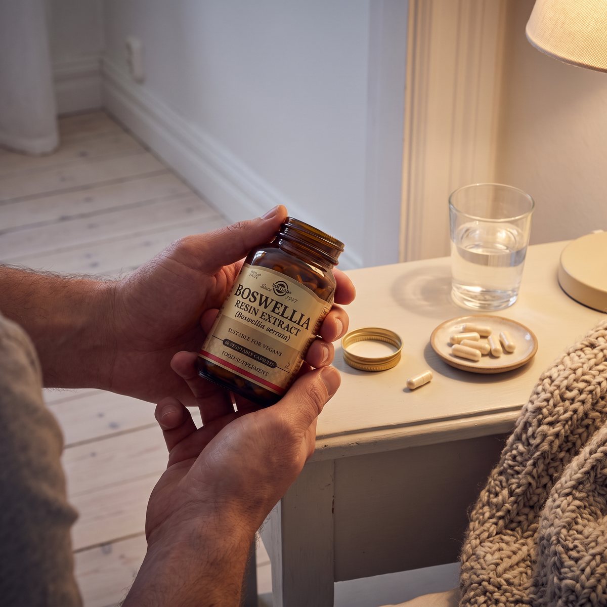 Solgar Boswellia-glasflaska i en hand vid ett nattduksbord med kapslar på ett fat och ett glas vatten i en svensk lägenhet