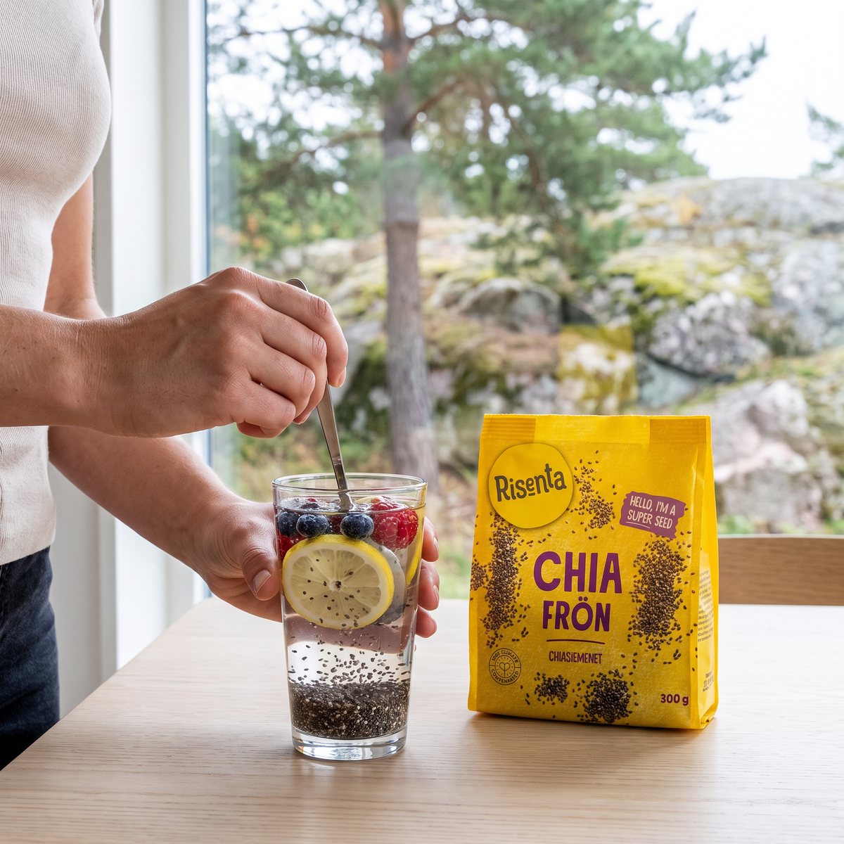 Gul påse med Risenta chiafrön bredvid ett glas vatten med frukt framför ett fönster med svensk tall och granit