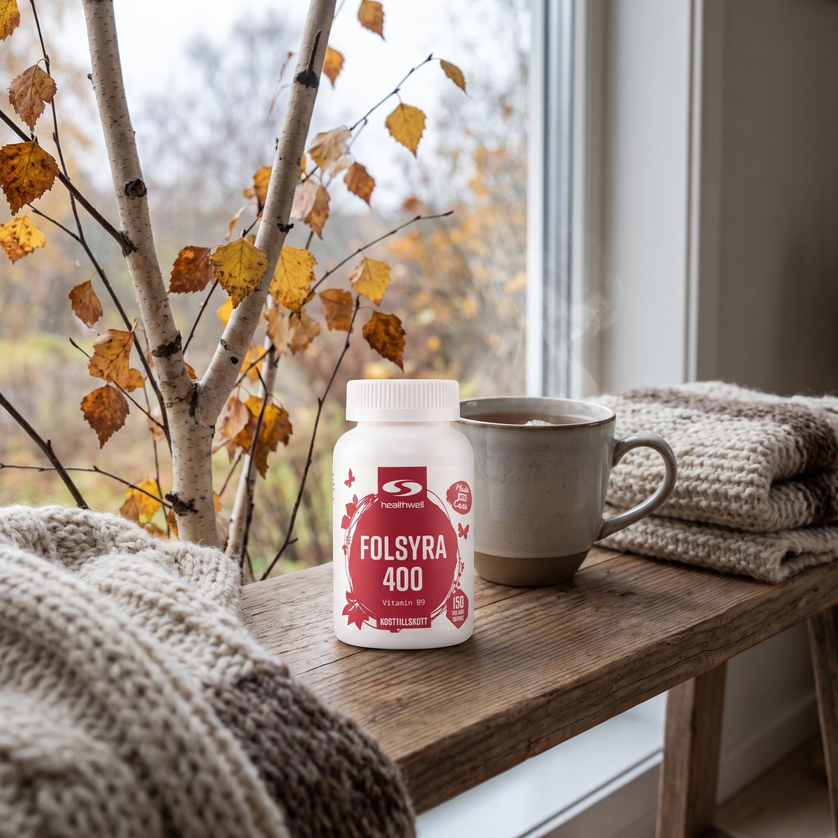 Healthwell Folsyra på en träbänk bredvid en tekopp och en stickad pläd en gråstilla svensk höstdag