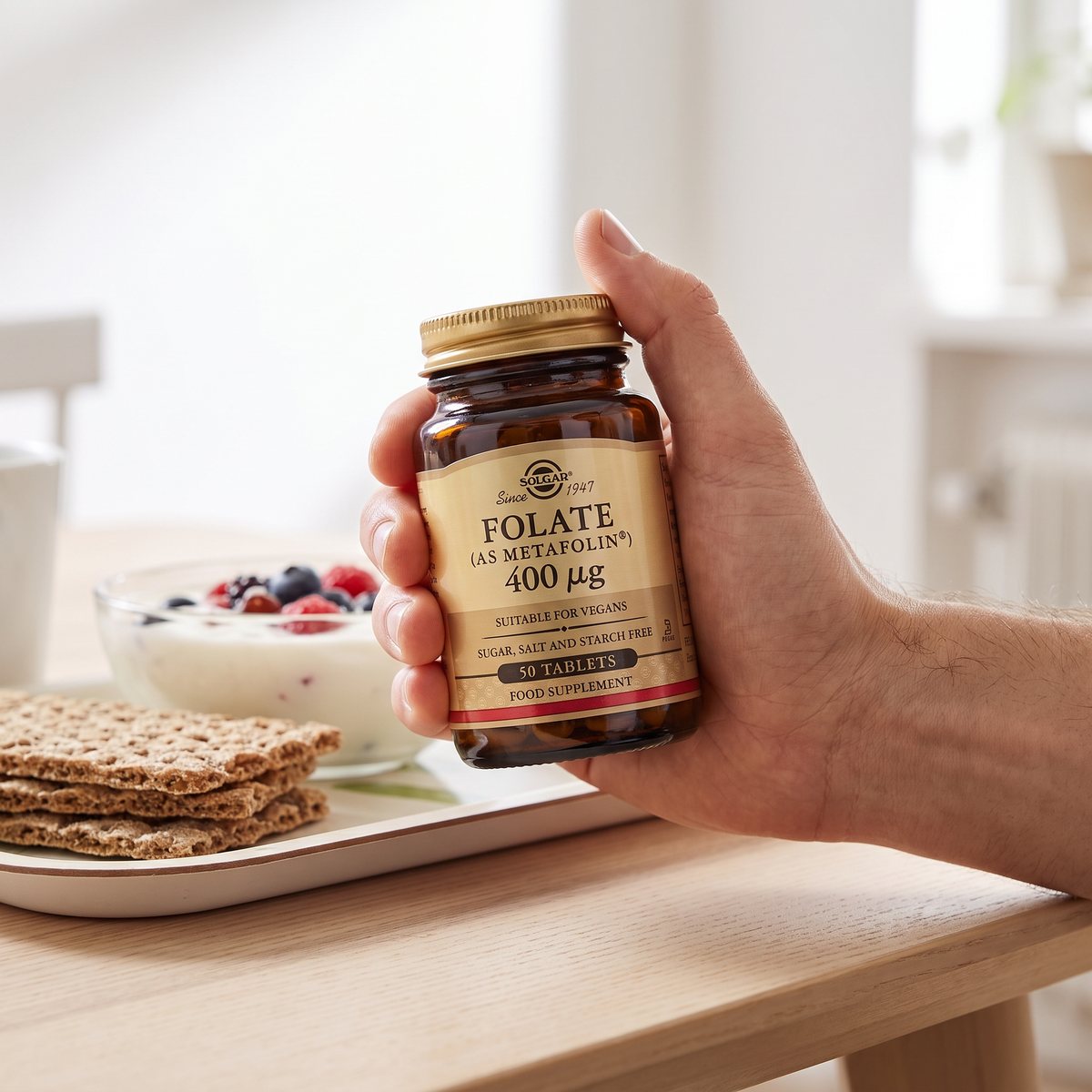 Hand som håller en burk Solgar Folat Metafolin vid ett frukostbord med yoghurt och knäckebröd i ett ljust svenskt hem