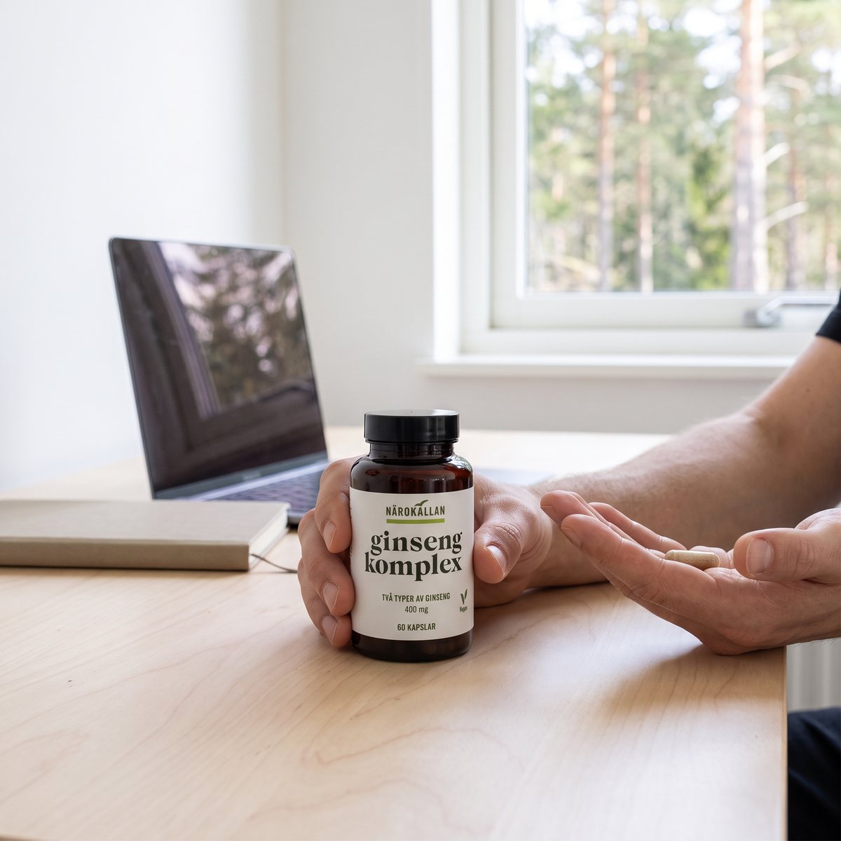 Närokällan Ginseng Komplex i brun glasburk hållen i handen vid ett skrivbord i ett svenskt hem