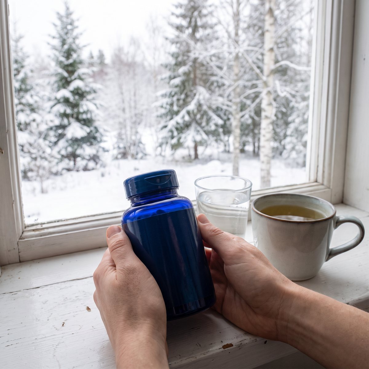Hand som håller Life Extension Mega Grönt Te Extrakt vid ett vitt snötäckt fönster i ett svenskt hem, med vatten och en kopp te