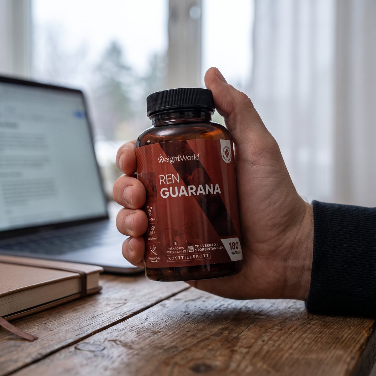 Brun burk med svart lock, WeightWorld Ren Guarana, i handen vid ett skrivbord i ett svenskt hemmakontor