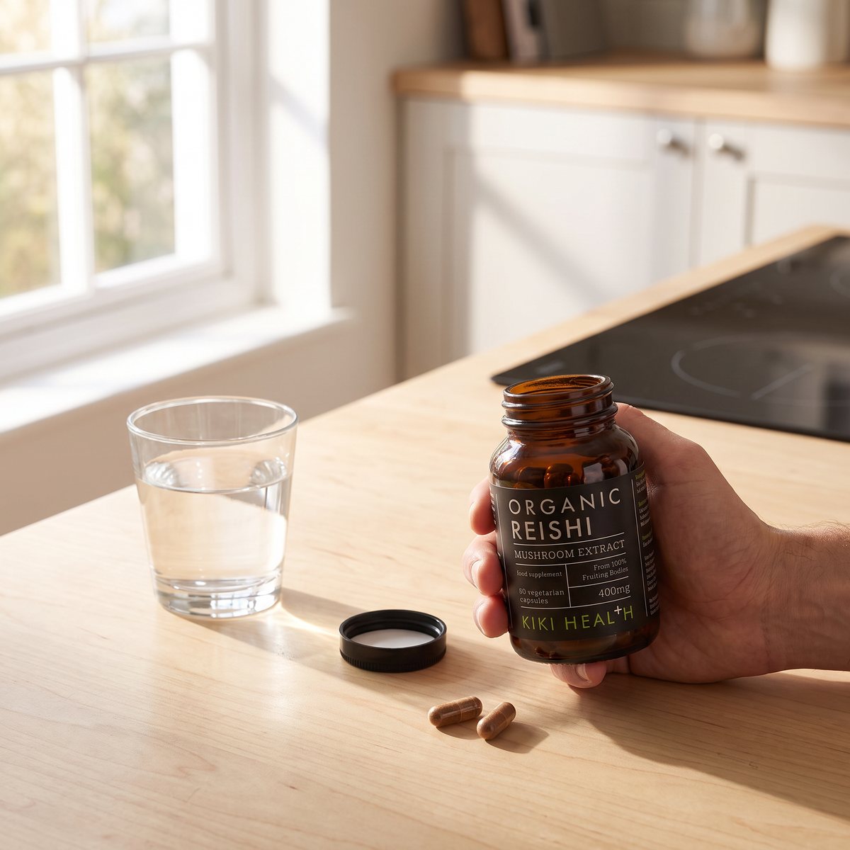 Brun glasburk med Kiki Health Organic Reishi hålls i handen vid ett glas vatten på ett ljust träbord i ett svenskt kök