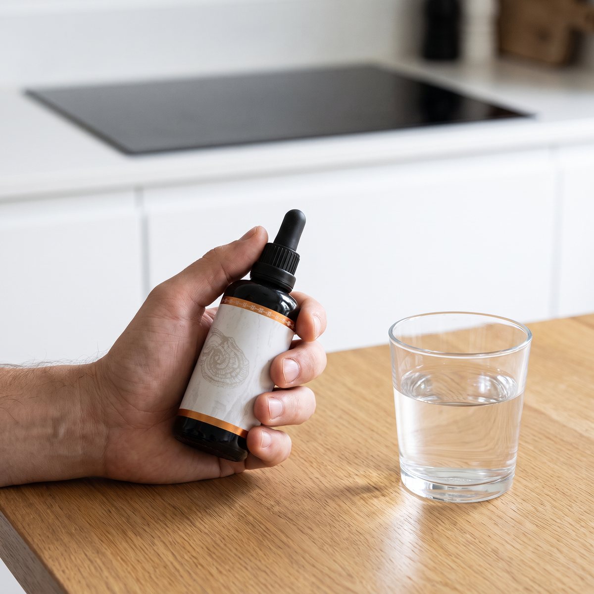 Hand håller en mörk pipettflaska med Rå Hygge Flytande Reishi Extrakt vid ett glas vatten på ett ekbord i ett svenskt kök