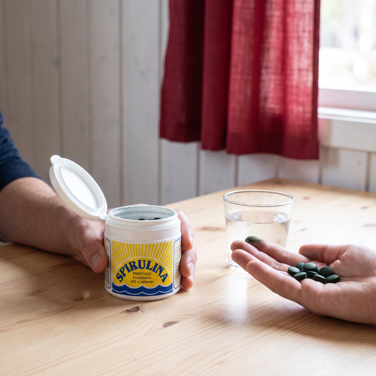 Lindroos spirulinaburk på ett vitt fönsterbräde i ett svenskt hem, med ett glas vatten och tabletter i handen.