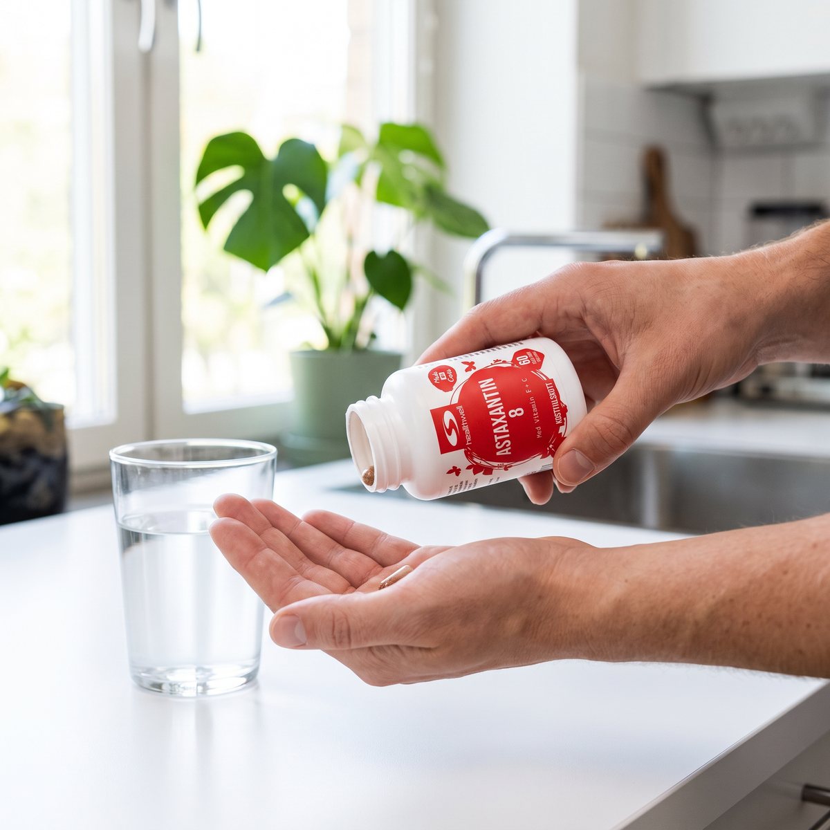 Vit burk med röd etikett, Healthwell Astaxantin, och en hand som vippar ut en kapsel vid ett vattenglas i ett svenskt kök