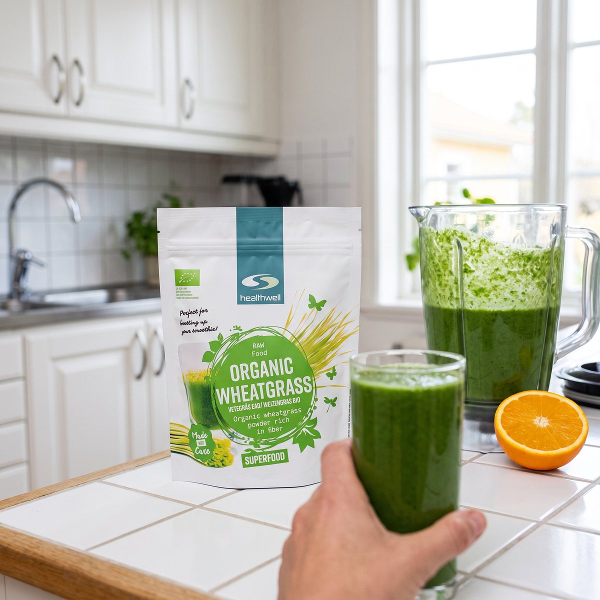 Healthwells vetegräspåse på en köksbänk bredvid en mixerkanna, en hand håller ett glas grön smoothie