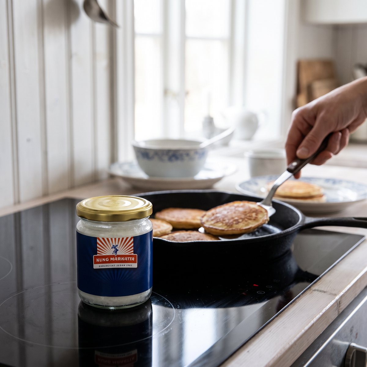 Liten glasburk med kokosolja bredvid en gjutjärnspanna med pannkakor på en induktionshäll i ett svenskt kök.