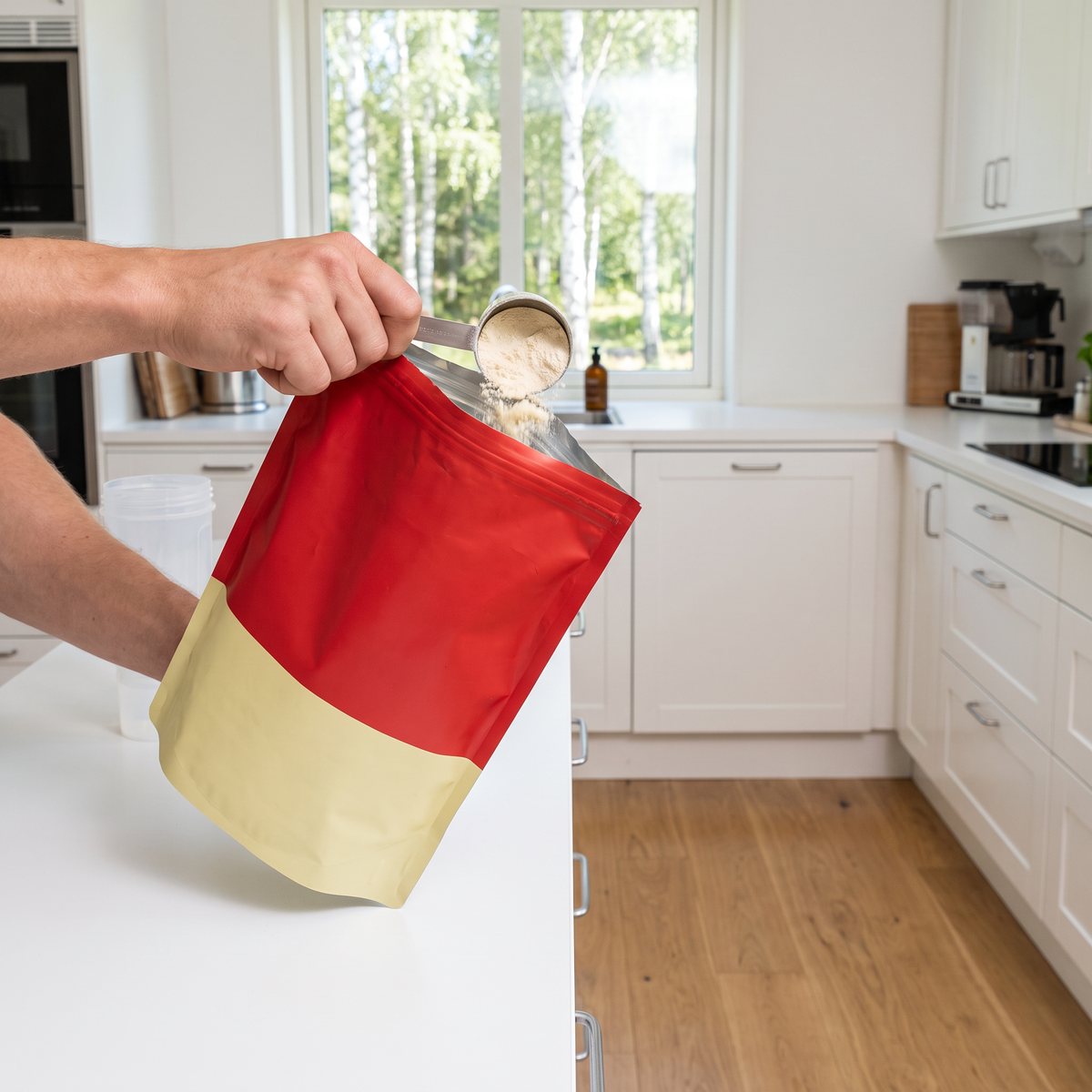 Tyngre Vassle Protein Vaniljdrömmar, röd och gul påse, som en hand häller pulver med mätsked i ett svenskt kök med vita luckor och trägolv