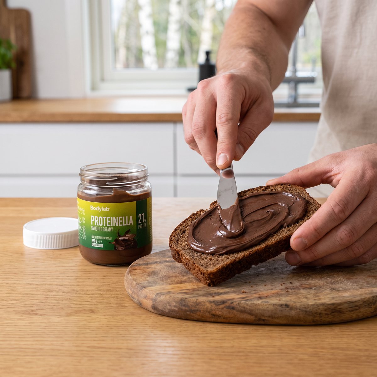 Burk med chokladproteinkräm med vitt lock bredvid en skiva rågbröd som bredas med krämen, på ett köksbord i ett svenskt kök
