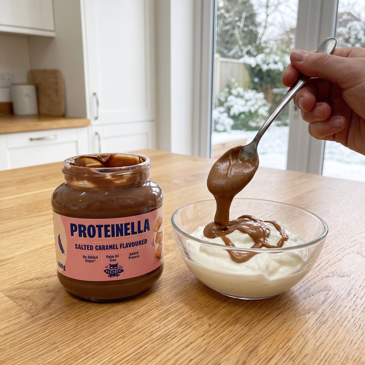 Burk med saltkaramellkräm bredvid en skål grekisk yoghurt där en sked fyller på krämen, i ett svenskt kök med snö utanför fönstret