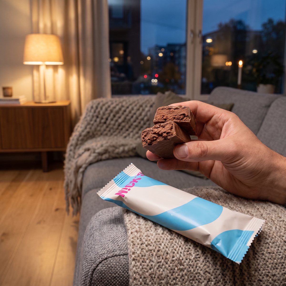 Nicks Protein Bar Triple Chocolate bruten i bitar i en hand ovanför en soffa med stickad pläd en höstkväll