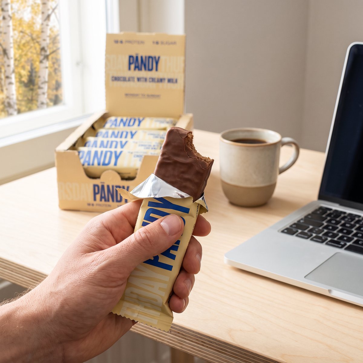 Pändy Protein Bar med halvt avskalat omslag i en hand vid ett ljust skrivbord med kaffemugg och laptop