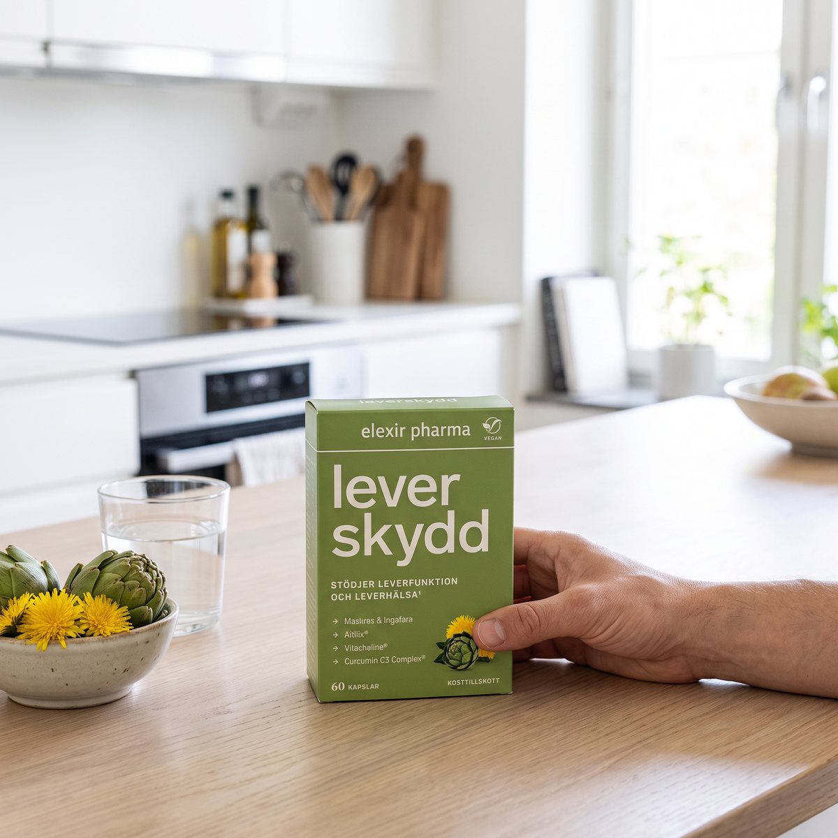 Förpackning med Elexir Pharma Leverskydd i handen på ett köksbord i ett svenskt kök, med vattenglas och kvistar av maskros och kronärtskocka