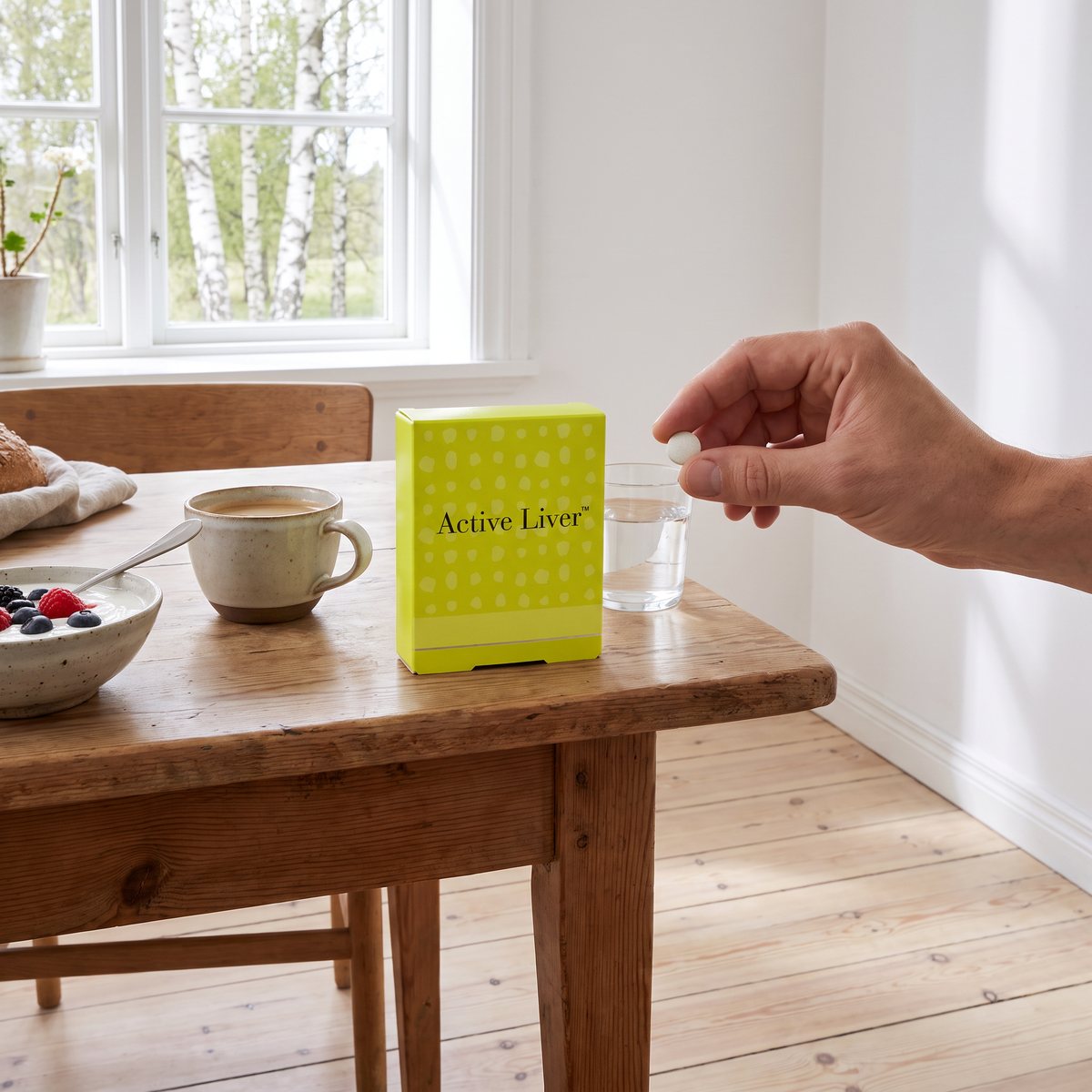 Ask med New Nordic Active Liver på ett träbord vid frukost i ett svenskt hem, med en hand som håller en tablett över ett vattenglas