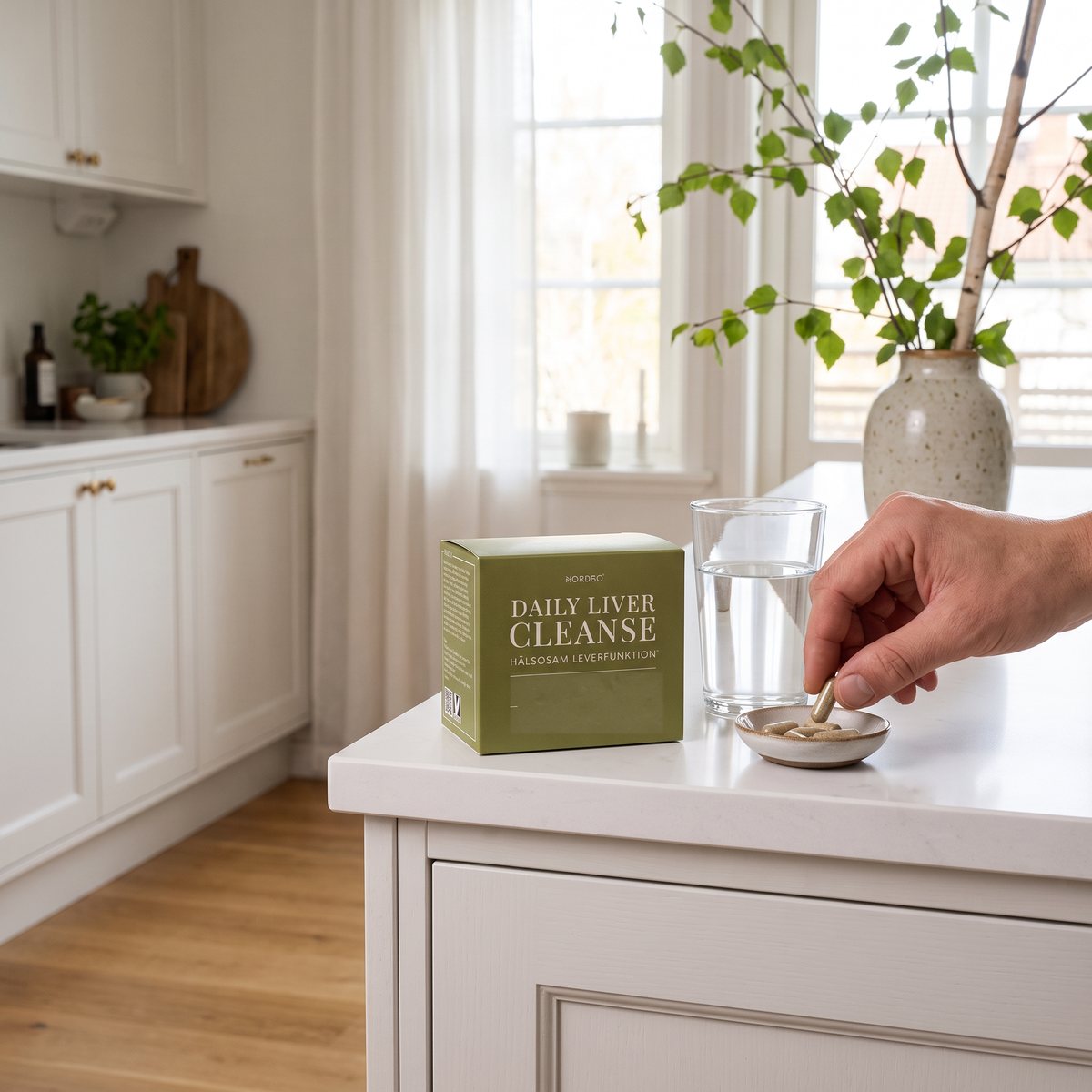 Kartong med Nordbo Daily Liver Cleanse på en vit köksbänk i ett svenskt kök, med vattenglas och en hand som tar en kapsel