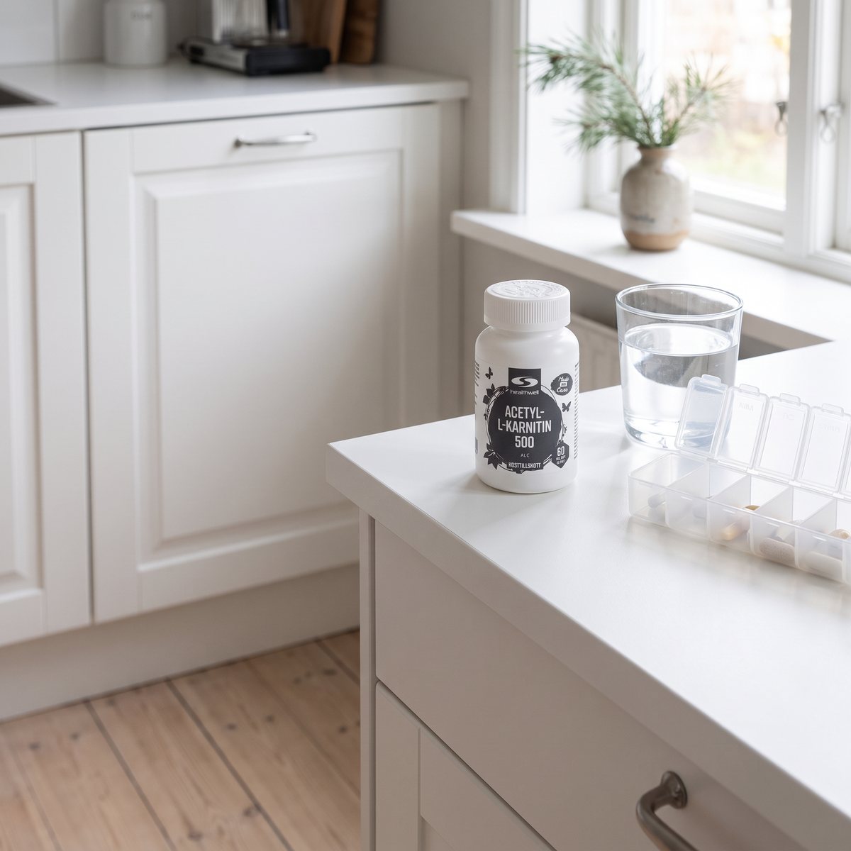 Healthwells vita burk Acetyl-L-karnitin på en vit köksbänk bredvid ett vattenglas och en pillerdosa i ett svenskt kök