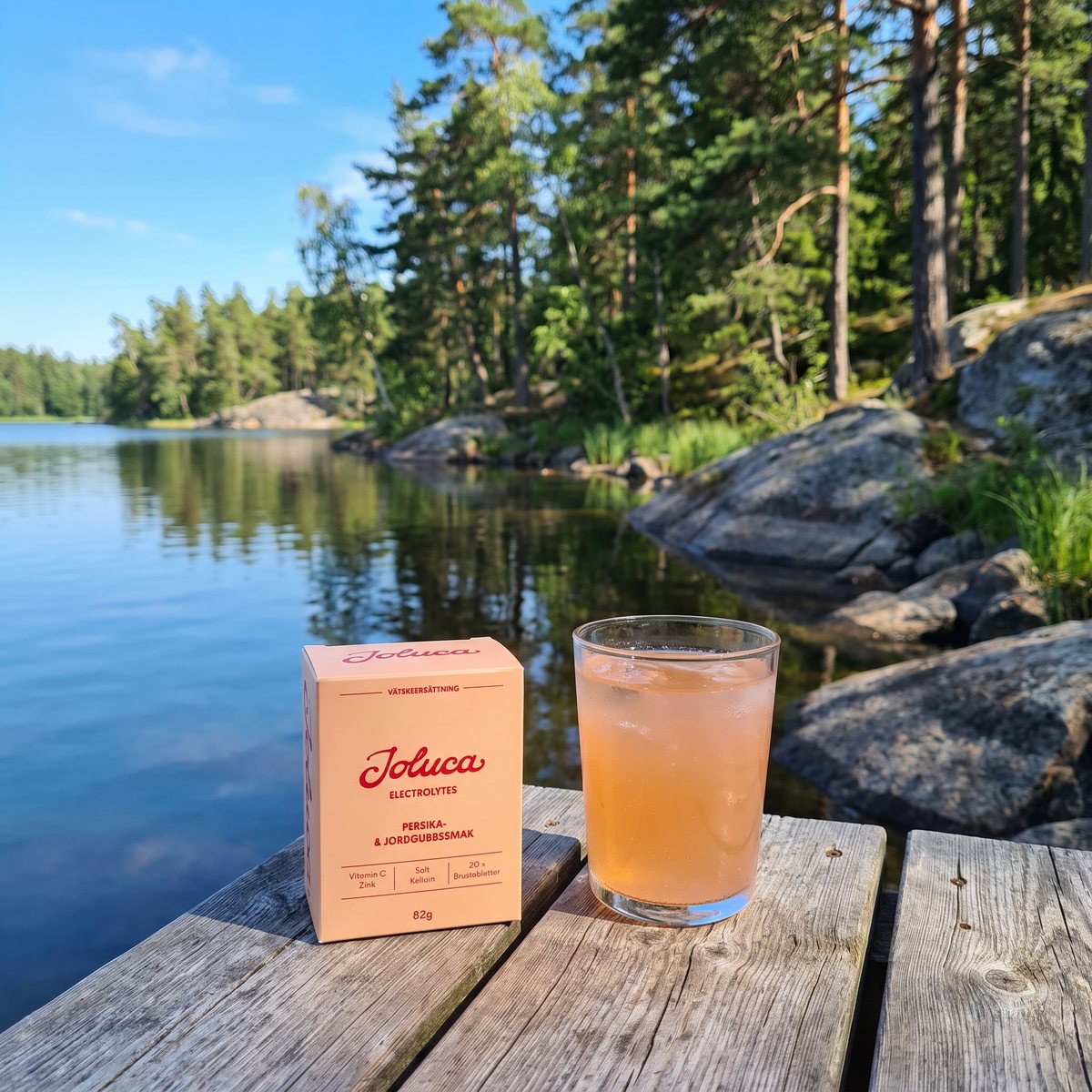 Joluca Rehydration Peach Strawberry: förpackningen på en träbrygga vid en svensk sjö med ett glas persikofärgad dryck.