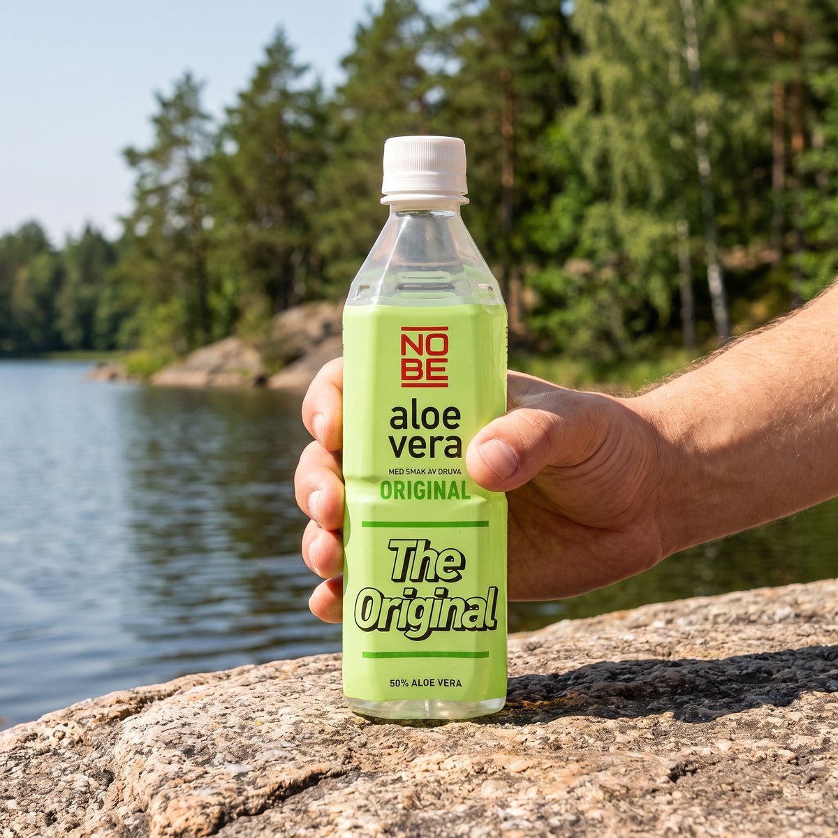 Nobe Aloe Vera Original, PET-flaska, hållen i handen på en granitklippa vid en svensk sjö