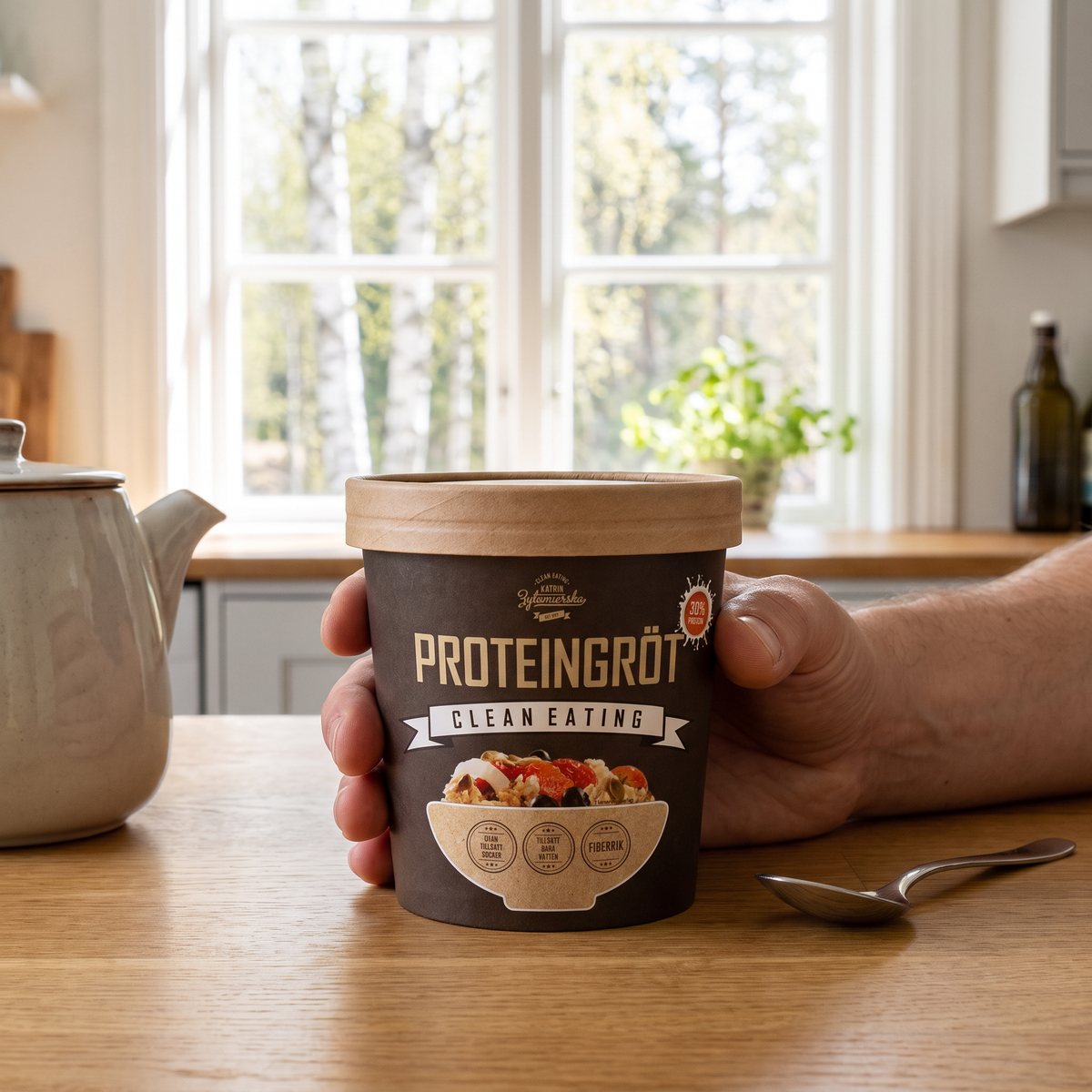 Clean Eating Proteingröt i pappersbägare hållen i handen vid ett fönster mot björkar i ett svenskt kök