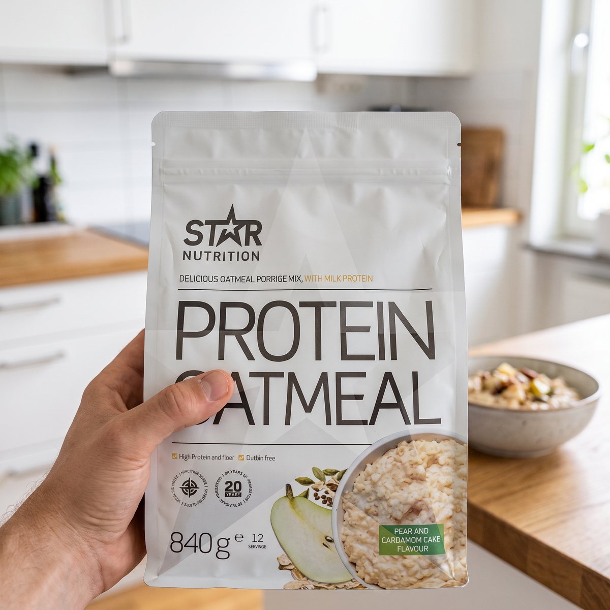 Förpackning med Star Nutrition Protein Oatmeal hållen i handen i ett ljust svenskt kök med en skål gröt i bakgrunden