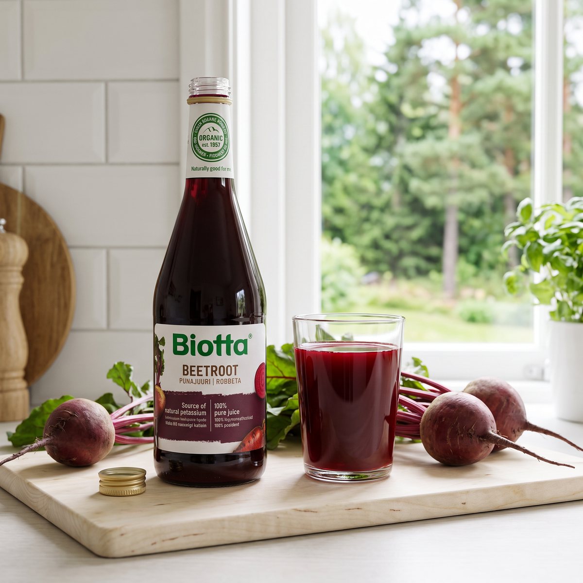 Biotta Rödbetsjuice på en skärbräda bredvid ett glas juice och färska rödbetor i ett svenskt kök med tallar utanför fönstret