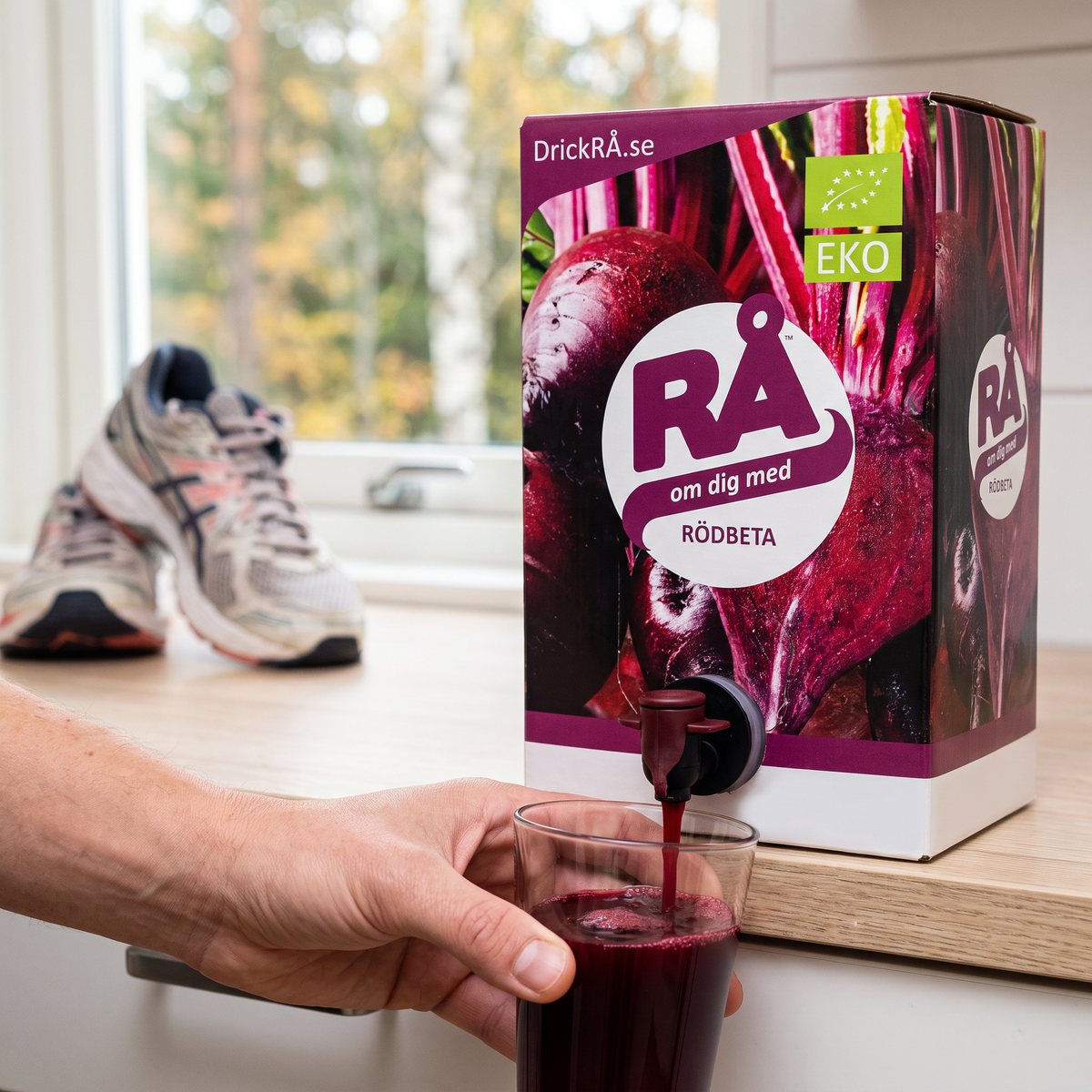RÅ Rödbeta i bag-in-box där rödbetsjuice tappas i ett glas på en köksbänk i en svensk lägenhet, med löparskor och höstträd i bakgrunden