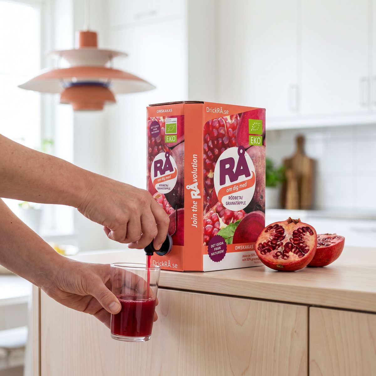 RÅ Rödbeta/Granatäpple i bag-in-box där juice tappas i ett glas i ett ljust svenskt kök, med ett delat granatäpple bredvid