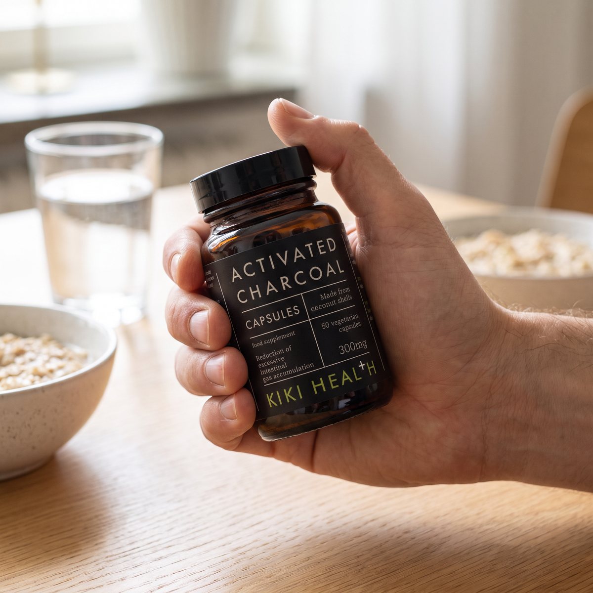 Hand som håller en brun glasburk med Kiki Health Activated Charcoal Caps vid ett frukostbord i ett svenskt hem