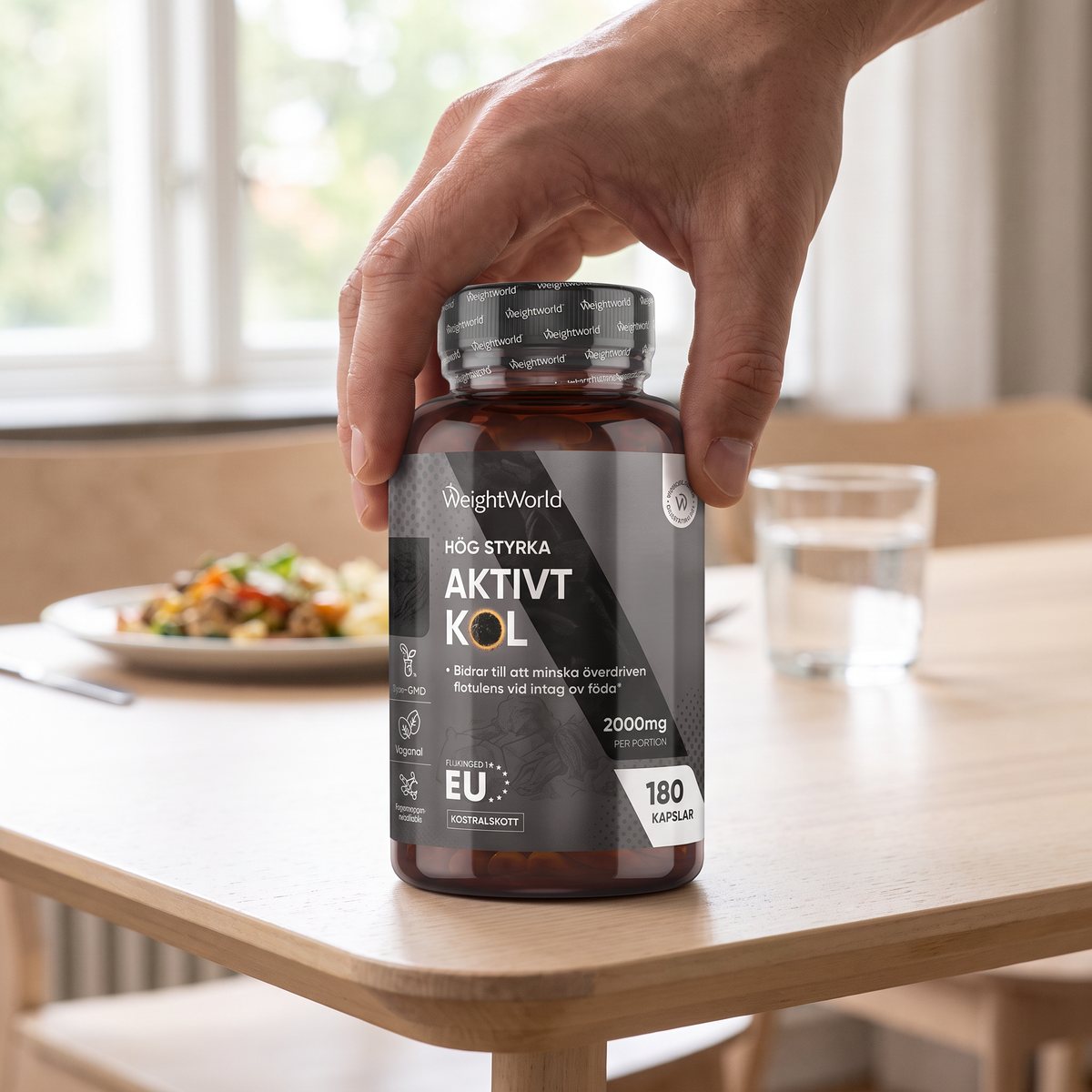 Hand som håller en burk WeightWorld Aktivt kol 2000 mg vid ett matbord i ett ljust svenskt hem