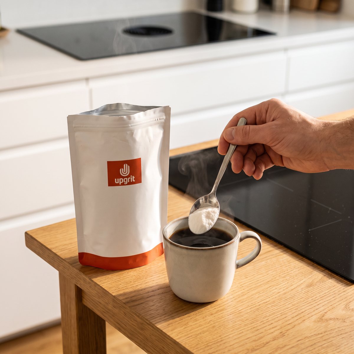 Upgrit C8 MCT-pulver i vit påse bredvid en kaffekopp där pulvret rörs i, på ett ljust ekbord i ett svenskt kök med induktionshäll