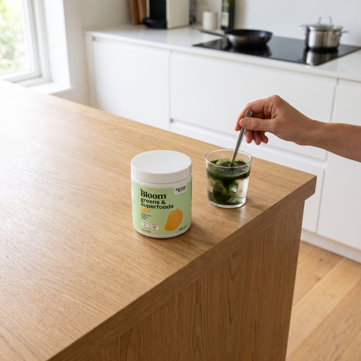 Burk med Bloom Greens & Superfoods på en köksö i trä medan en hand rör ut pulvret i ett glas vatten i ett svenskt kök