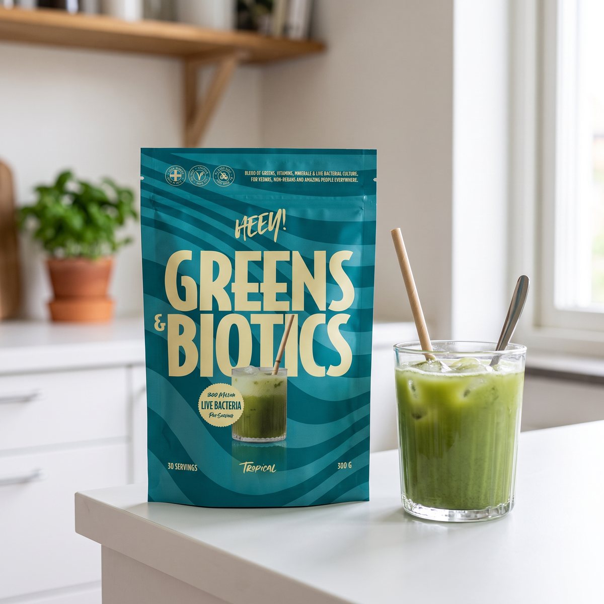 Påse med Heey Greens & Biotics Tropical bredvid ett glas grön iskall dryck med sugrör på en köksbänk i en svensk lägenhet