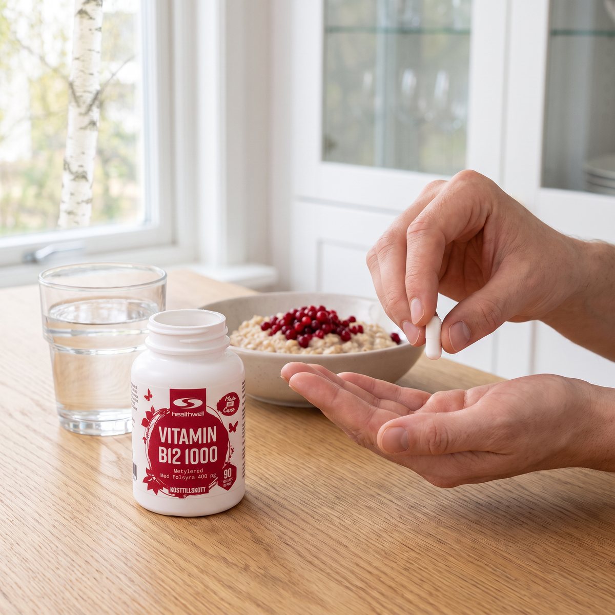 Burk Healthwell Vitamin B12 1000 Metylerad på ett köksbord i ek bredvid ett vattenglas och havregrynsgröt med lingon, en hand håller en kapsel