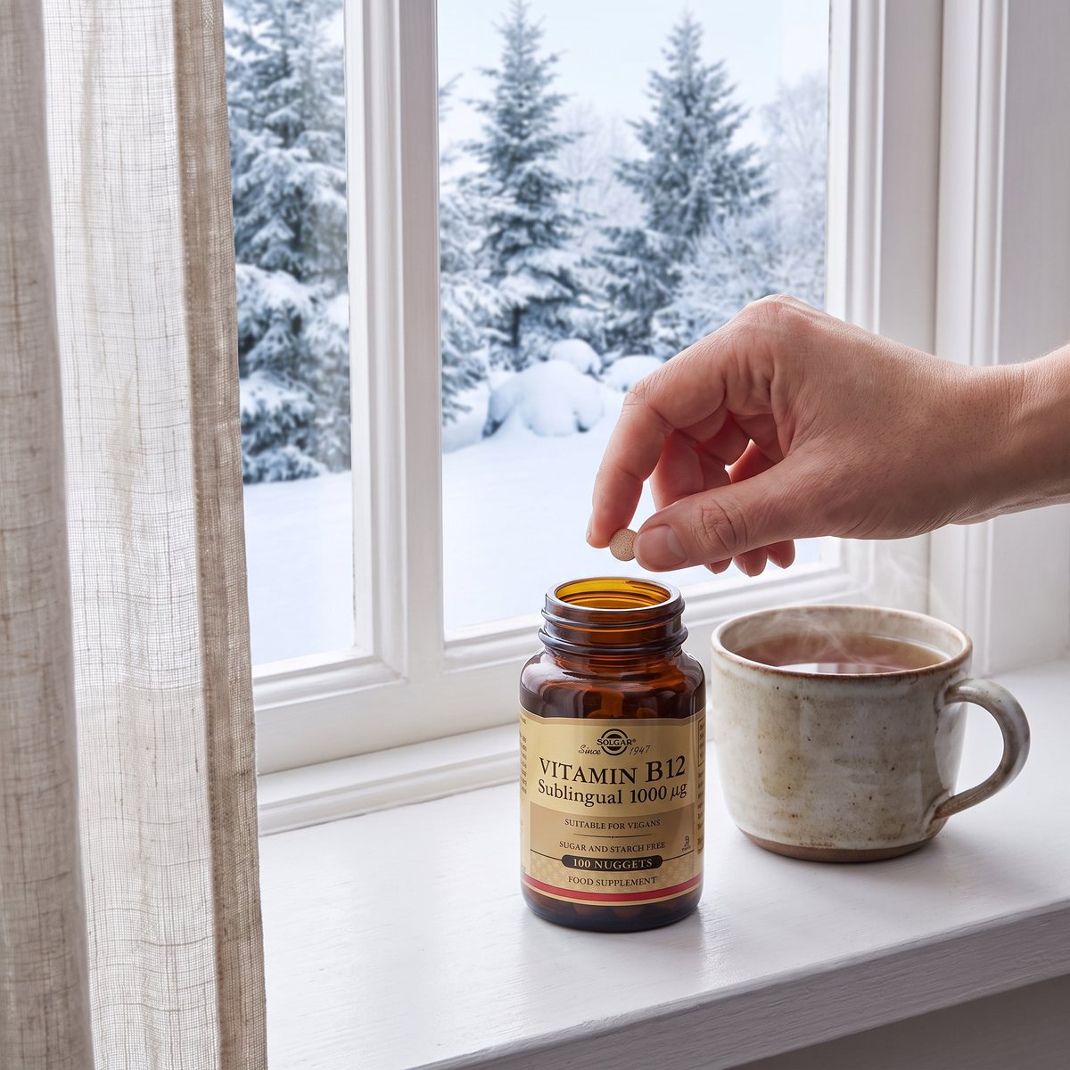 Brun glasburk Solgar Vitamin B12 1000 mcg på en vit fönsterbräda bredvid en tekopp, en hand tar en sugtablett, snöig granträdgård utanför