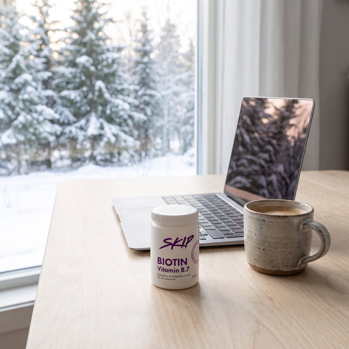 Vit burk med Skip Biotin vid en bärbar dator och en kaffekopp på ett skrivbord i ett svenskt hem med snöig granskog utanför