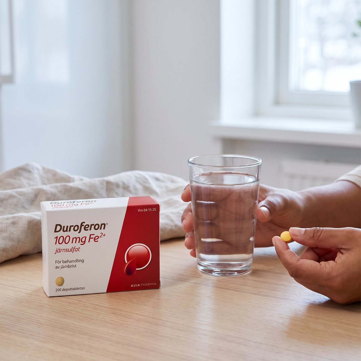 Förpackning med Duroferon 100 mg på ett köksbord bredvid ett glas vatten, en hand håller en liten gul rund tablett