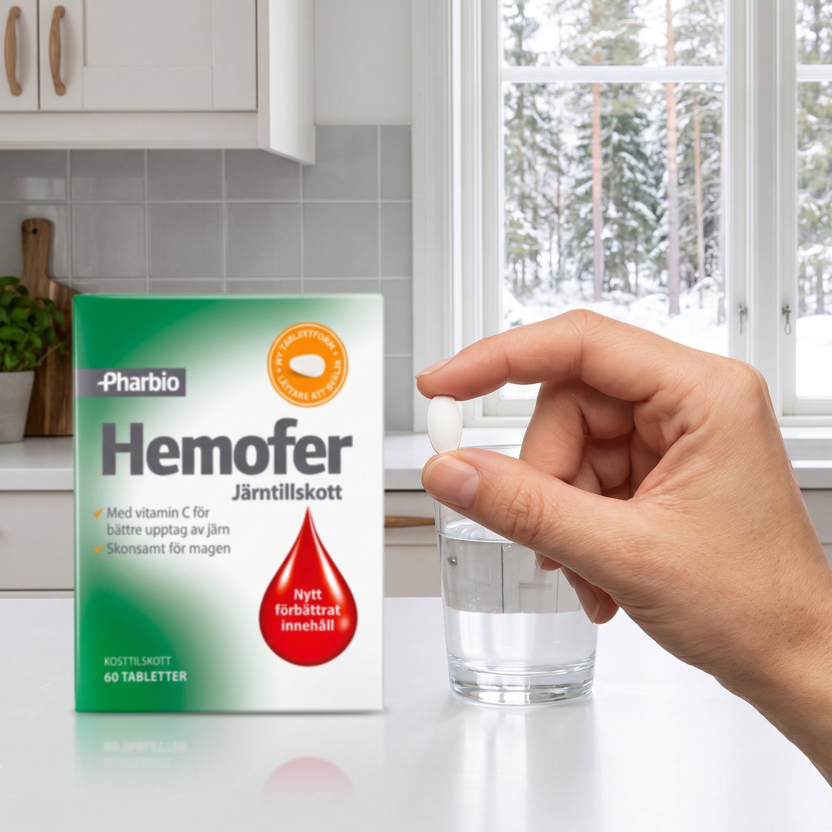 Förpackning med Pharbio Hemofer på en vit köksbänk bredvid ett glas vatten, en hand håller en vit tablett framför ett fönster mot snöig granskog