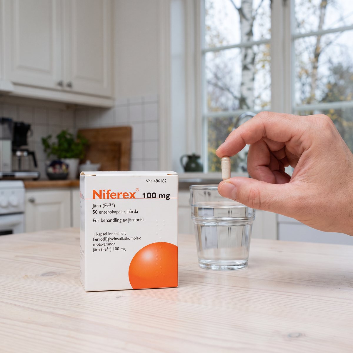 Förpackning med Niferex 100 mg på ett köksbord i trä bredvid ett glas vatten, en hand håller en kapsel framför ett fönster med björkar